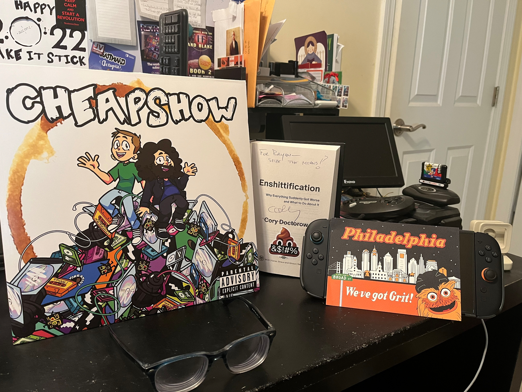 A cluttered desk with five items set up on display. From left to right: The CheapShow Vinyl record leaning against my M-Stand, a pair of glasses, a signed copy of Cory Doctorow’s latest book, Enshittification, with a swearing poop emoji sticker now stuck properly on the page underneath his signature, a Nintendo Switch 2 console propped up by its kickstand, and a postcard depicting the Philadelphia skyline with the city’s fuzzy mascot, Grit, popping up from the bottom-right corner. 