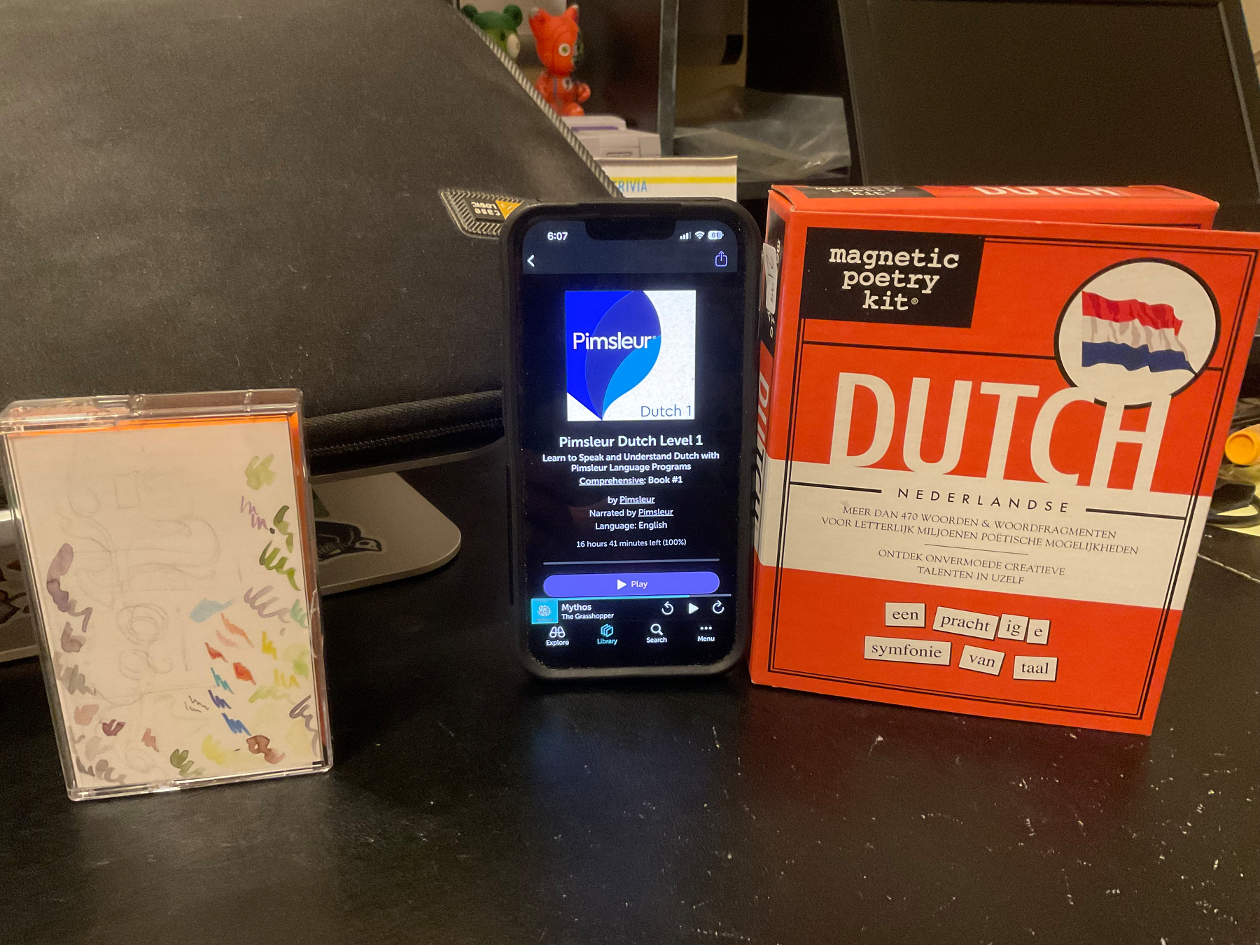 My desk with three things in front of my laptop stand. From left to right: A cassette tape from Pocket Vinyl titled “Banned Books”, my iPhone 13 displaying the UI for Libro.fm with Pimsleur’s Dutch Level 1 on display, and an orange box from Magnetic Poetry Kit containing Dutch words to create sentences with.